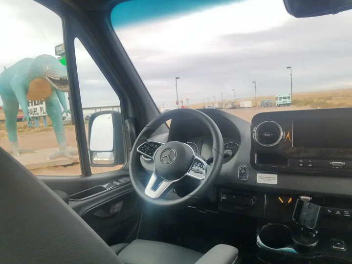 RV rental van interior overlooking a large blue dinosaur statue at a travel plaza with other parked RVs, perfect for travel.. Winnebago Revel 2020