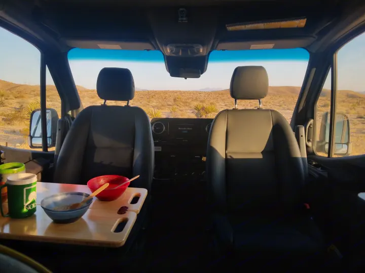 Inside an **rv rental** parked in the desert, two bowls and a mug sit on a table, with open views of sandy terrain and mountains.. Winnebago Revel 2020