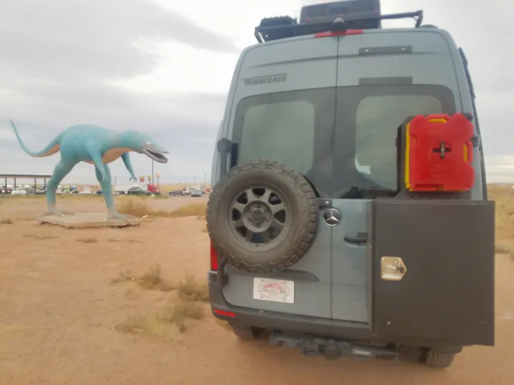 This rv rental Winnebago camper van with spare tire, storage & fuel sits by a large blue dinosaur statue. Roadside attraction.. Winnebago Revel 2020