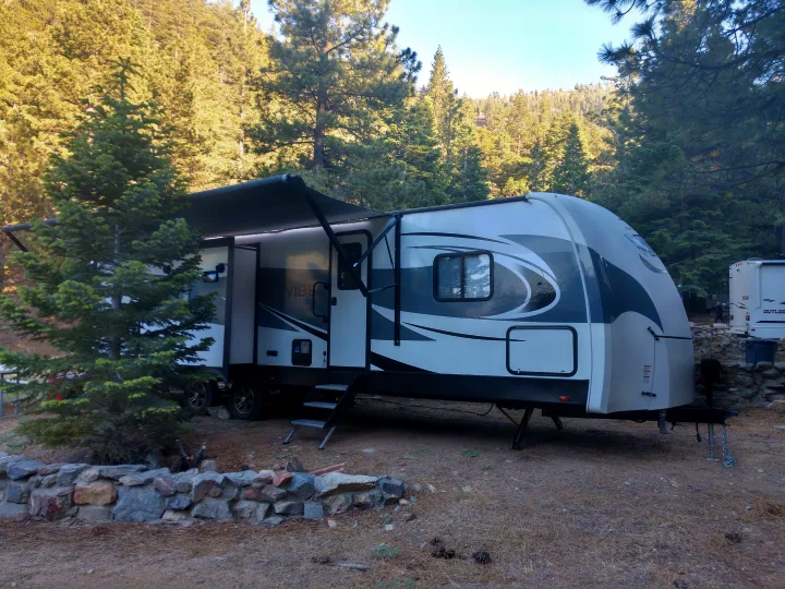 Modern travel trailer rv rental set up with awning extended at a wooded campsite, surrounded by tall pine trees.. Forest River Vibe 2018