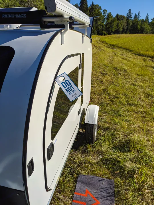 A white teardrop RV rental with a 'Do Not Disturb' sign on its window, parked in a sunny, grassy field.. DROPLET 58 2019