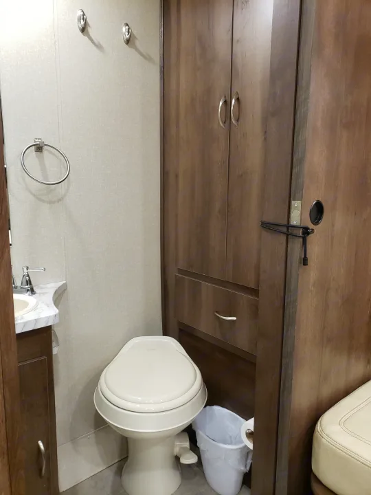 Inside an RV rental bathroom, featuring a compact toilet, sink, storage cabinets, towel ring, hooks, and trash can. Functional for travel.. Coachmen Other 2018