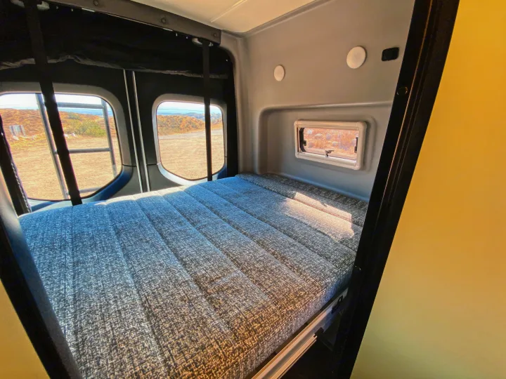 Inside an RV rental: a comfortable bed with a textured gray mattress, flanked by scenic rear windows overlooking a dry landscape.. Winnebago Revel 2020