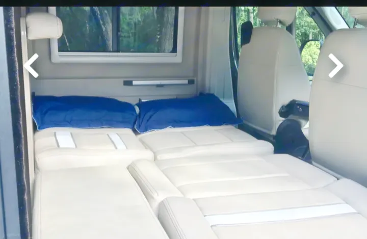 Inside an RV rental: a comfortable beige bed with blue pillows, extending from the rear toward swiveled front seats, ready for travel.. Winnebago Travato 2019