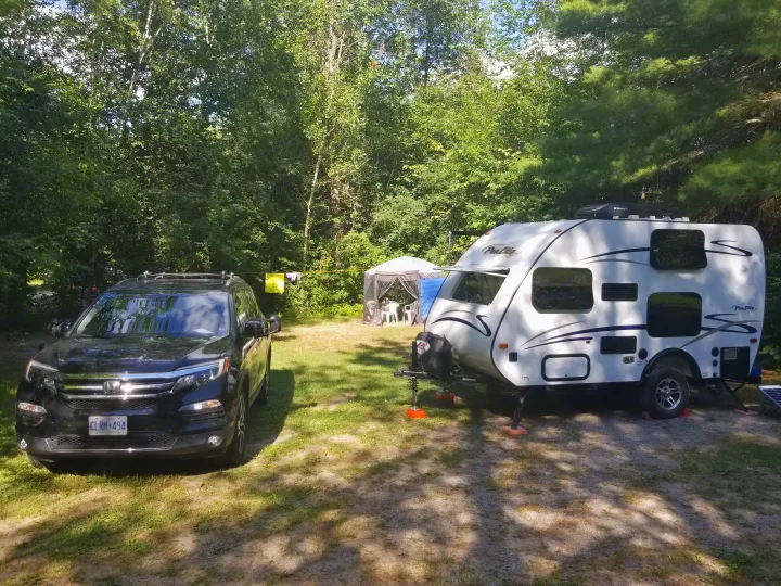 An RV rental setup: a black SUV and white travel trailer parked at a sunny, wooded campsite with a screen tent.. Prolite Prolite Trailer 2015