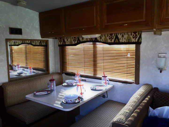 Inside an RV rental: a cozy dinette booth with patterned seating, a set table, wooden cabinets, and a window. Perfect for camper meals.. Fleetwood Bounder 1999