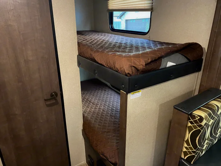 View inside an RV rental showing two neatly made bunk beds with brown quilted bedding. A window provides an outdoor glimpse.. Other Sun-Lite 2019