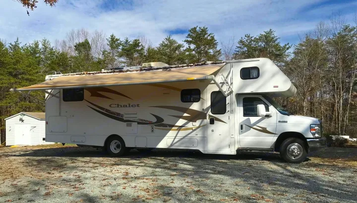 White Class C RV rental with extended awning, parked on gravel in a wooded area. Perfect for your outdoor escape.. Winnebago Chalet 2014