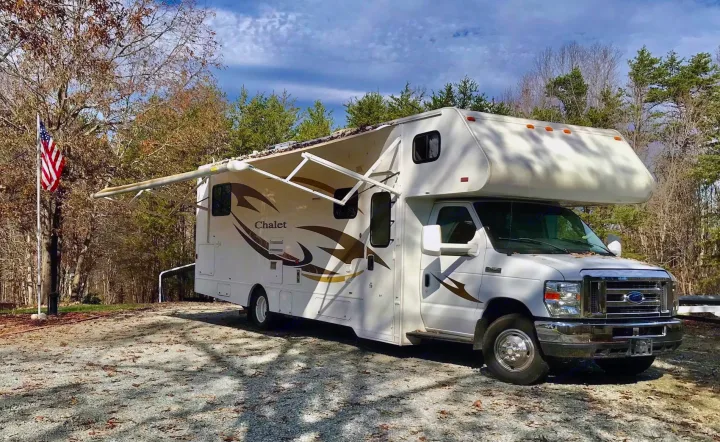 White Chalet RV rental parked in an outdoor setting with an extended awning. Trees and an American flag complete the scene.. Winnebago Chalet 2014