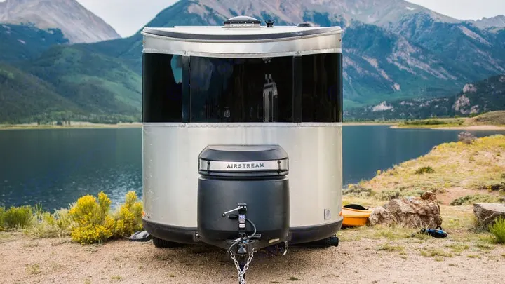 A sleek Airstream RV rental rests by a lake, surrounded by mountains.. Airstream Basecamp 2023