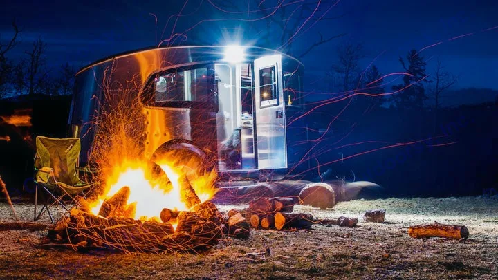 Cozy nighttime rv rental camping scene: a bright campfire with sparks, a chair, and a shining Airstream under dark skies.. Airstream Basecamp 2023