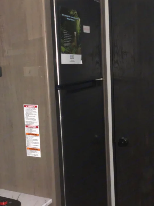 An RV rental's compact interior features a black refrigerator/freezer, with prominent safety warnings on the adjacent wall.. Keystone Hideout 179LHS 2021