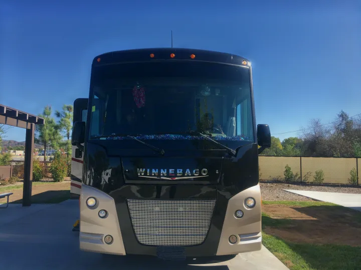 Front of a large black and tan Winnebago motorhome, an rv rental, parked on a sunny day at a campsite.. Winnebago Vista 2016