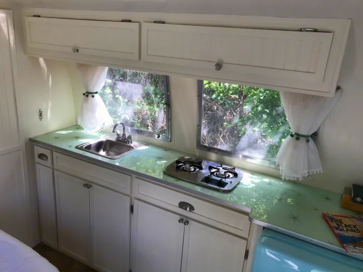 Step into this charming retro RV rental kitchen with green starburst counters, white cabinets, sink, & stove. Perfect vintage camper interior.. Airstream Caravel 1968