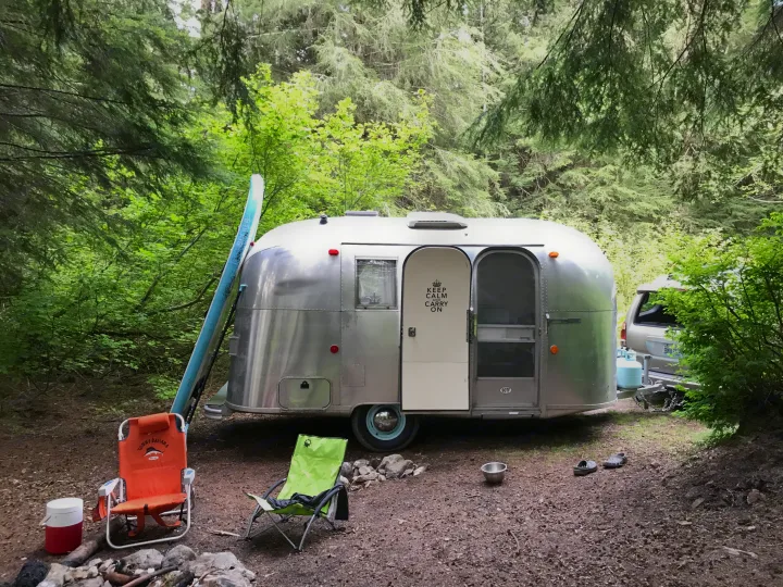 A scenic RV rental setup featuring a polished Airstream trailer, camping chairs, and a paddleboard at a shaded campsite.. Airstream Caravel 1968