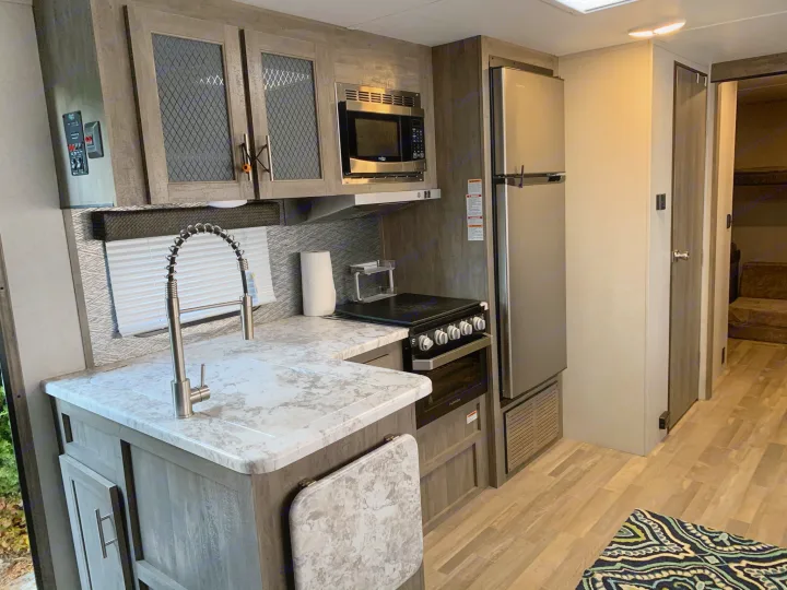 Modern RV rental kitchen: light wood-look cabinets, white counters, sink, stainless steel microwave, fridge, and oven.. Forest River Vibe 2021