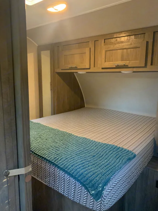 RV rental interior: well-lit bedroom with bed, herringbone mattress, teal blanket, and wood cabinets.. Forest River Vibe 2021