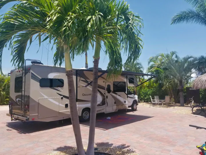 A beige motorhome RV rental is parked on a sunny paver driveway, surrounded by lush palm trees, perfect for a tropical getaway.. Thor Quantum 2017