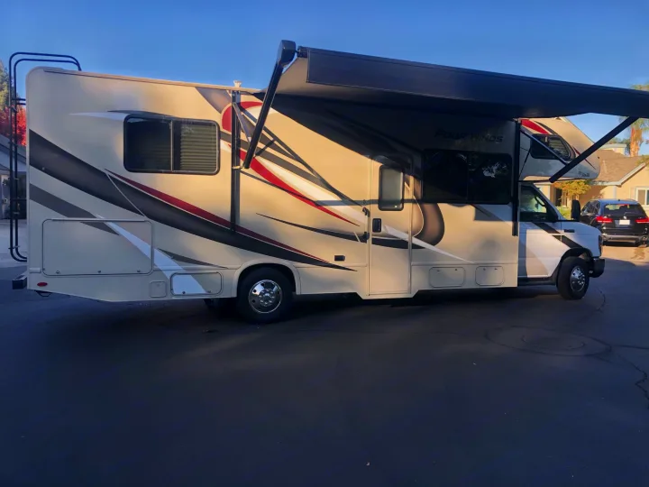An RV rental parked on asphalt, featuring a beige Four Winds motorhome with dark/red graphics and an extended awning.. Thor Four Winds 2020