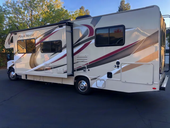 This spacious, beige RV rental motorhome with its slide-out open is parked on asphalt, ready for travel adventures.. Thor Four Winds 2020