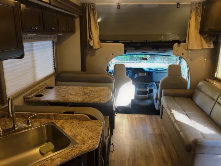 Interior view of a modern **rv rental** with kitchen, dining booth, sofa, and front cabin. Features light finishes and wood flooring.. Thor Four Winds 2020