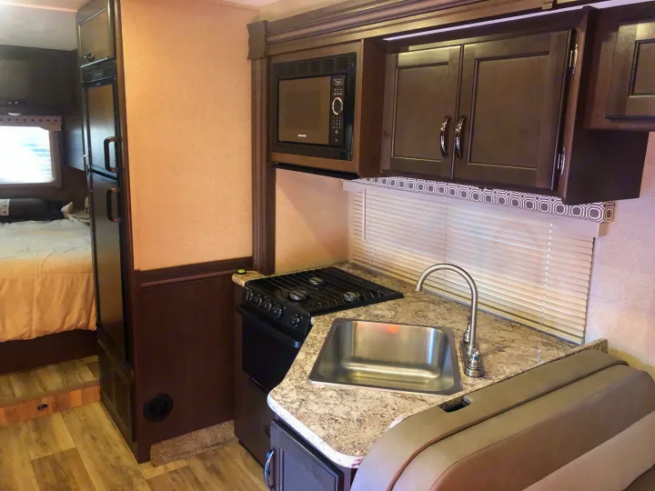 RV rental interior showing a compact kitchen with stove, sink, microwave, and adjacent bedroom area.. Thor Four Winds 2020