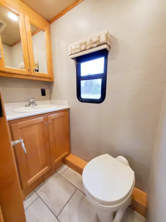 Inside an RV rental bathroom: light wood vanity, sink, mirrored cabinet, small window, and white toilet on tiled floor.. Tiffin Motorhomes Allegro Red 2021