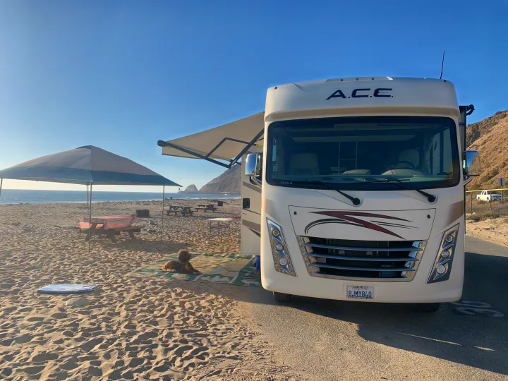 An RV rental parked at a sunny beach campsite, complete with a dog, picnic setup, and ocean view.. Thor Motor Coach A.C.E 2020