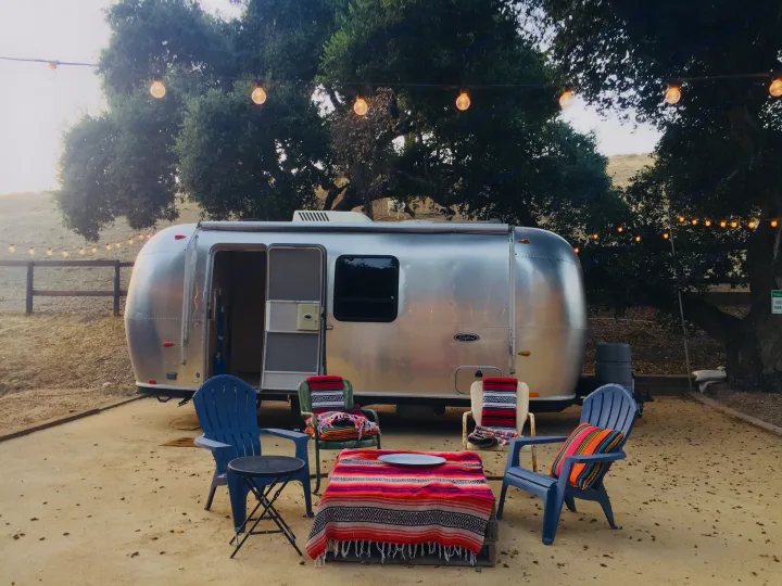 A silver Airstream rv rental at dusk, with outdoor seating & string lights creating a cozy glamping spot.. Airstream Safari 2008