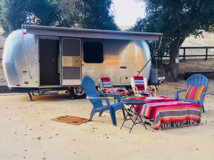 Cozy Airstream RV rental setup at a scenic campsite, featuring vibrant outdoor living with chairs and a blanket-covered table.. Airstream Safari 2008
