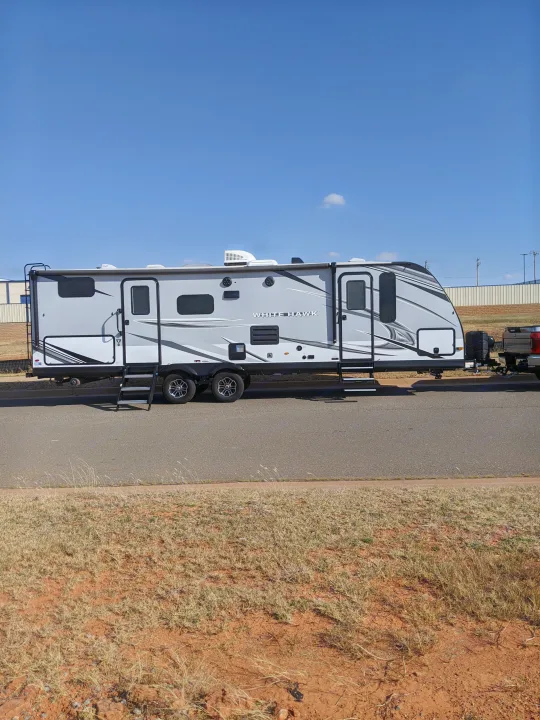 A large White Hawk travel trailer, an RV rental, is parked on asphalt under a clear blue sky, hitched to a pickup truck.. Jayco White Hawk 2021