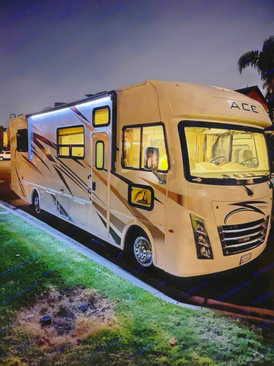 This night shot shows an illuminated RV rental parked on a street, glowing brightly inside with cool blue awning lights.. Thor Motor Coach A.C.E 2020