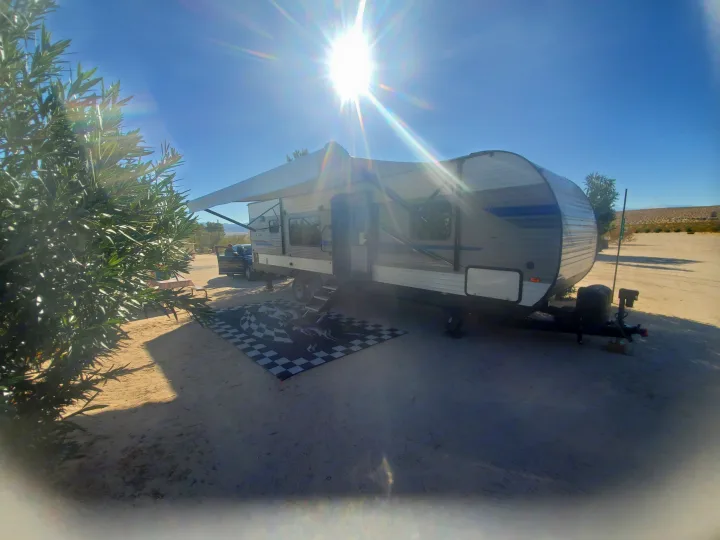 Sunny desert RV rental campsite featuring a travel trailer with extended awning & checkered mat, plus a parked pickup.. K-Z Manufacturing Sportsmen 2019