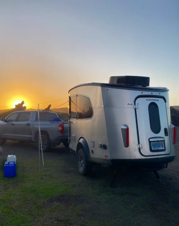 A silver Airstream travel trailer is hitched to a pickup truck at sunset, suggesting an ideal RV rental. Camping gear visible.. Airstream Basecamp 2023