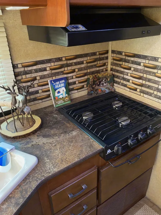 An RV rental kitchen featuring a three-burner stovetop, exhaust fan, decorative backsplash, and an "RV Cooking Made Easy" cookbook.. Jayco Redhawk 2014