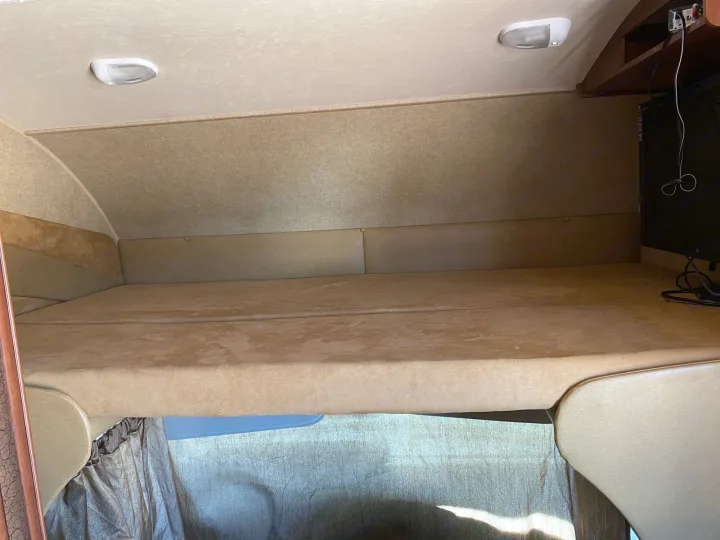 This image shows a cozy overhead sleeping bunk in an RV rental, featuring a comfortable mattress, ceiling light, and TV setup.. Jayco Redhawk 2014