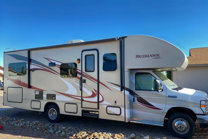 Jayco Redhawk Class C motorhome RV rental, tan with red & brown graphics, parked on gravel under a clear blue sky.. Jayco Redhawk 2014