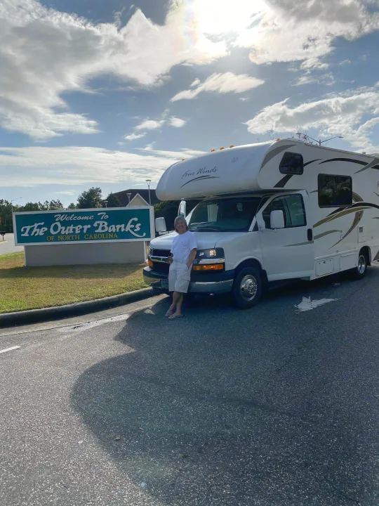 Excited traveler posing with their RV rental at the 'Welcome to The Outer Banks of North Carolina' sign on a sunny day.. Thor Motor Coach Four Winds 2013