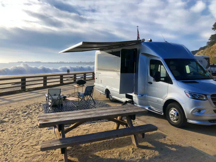 Coastal rv rental setup: silver RV with awning, picnic table, and chairs by crashing ocean waves. People watch the sea.. Airstream Atlas 2021