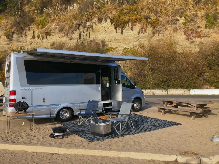 Coastal RV rental setup with Airstream camper van, awning, chairs, cooler & picnic table on sand near cliff.. Airstream Atlas 2021