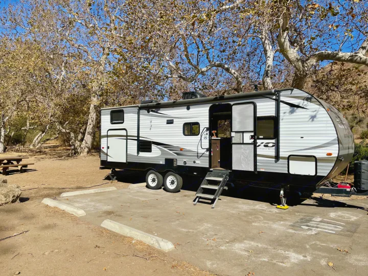 White & gray RV rental (travel trailer) parked at a campsite with a picnic table, under trees with sparse leaves. Ideal for camping.. Forest River Evo T2550 2019