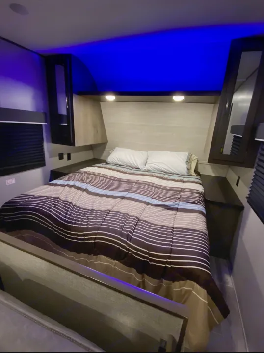 Cozy RV rental bedroom with a striped comforter on the bed, accented by bright blue overhead lighting and built-in storage.. Jayco Jay Flight SLX 2021