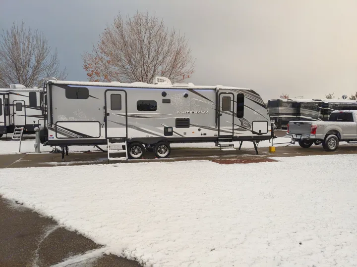 A snow-dusted RV rental travel trailer is parked with a pickup truck in a winter campground. Bare trees are visible under a grey sky.. Jayco White Hawk 2021