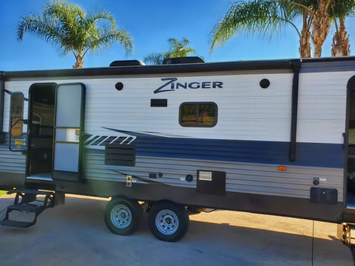 A clean Zinger travel trailer RV rental with open doors and steps, parked on pavement under a bright blue sky with palm trees.. Crossroads Zinger 2018