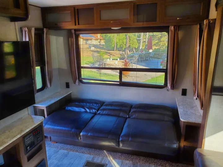 Inside an **RV rental**: a brown leather sofa, TV, and large window overlooking an outdoor patio scene.. Crossroads Zinger 2018