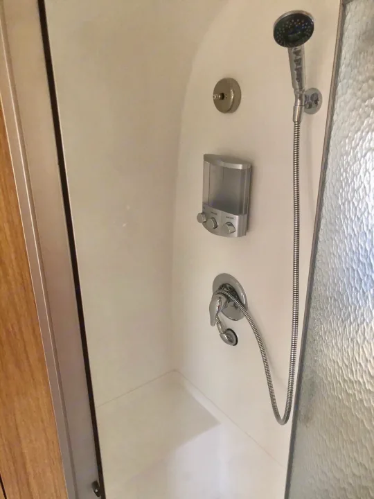 Compact RV rental shower: White-walled stall with a handheld shower, wall-mounted soap dispenser, and textured glass door.. Airstream Flying Cloud 2019