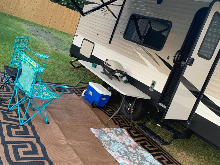 An RV rental campsite features a travel trailer with awning, grill, cooler, and colorful chairs set on an outdoor mat for relaxing.. Keystone Hideout 2019