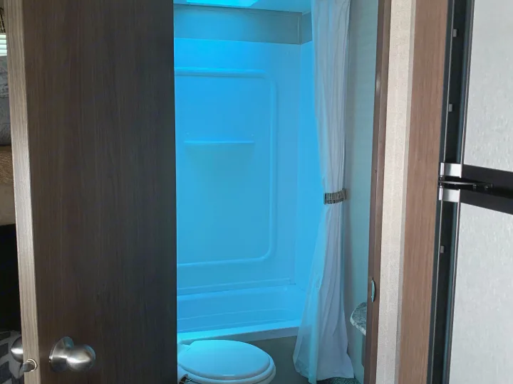 View into an RV rental bathroom with a blue-glowing shower/tub, toilet, and shower curtain.. Keystone Hideout 2019
