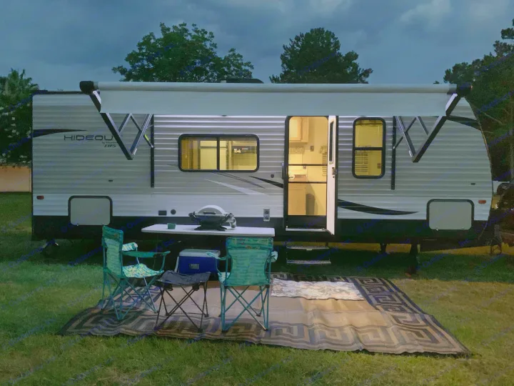 Cozy RV rental setup at night: illuminated awning, open door, grill, chairs, and outdoor mat on grass.. Keystone Hideout 2019