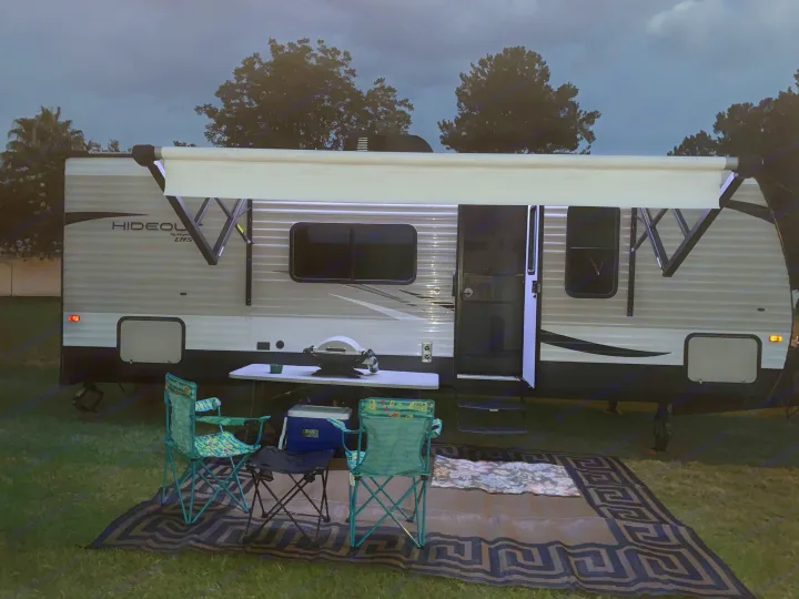 Nighttime RV rental setup: travel trailer with awning, chairs, grill on mat. Perfect for outdoor camping leisure.. Keystone Hideout 2019
